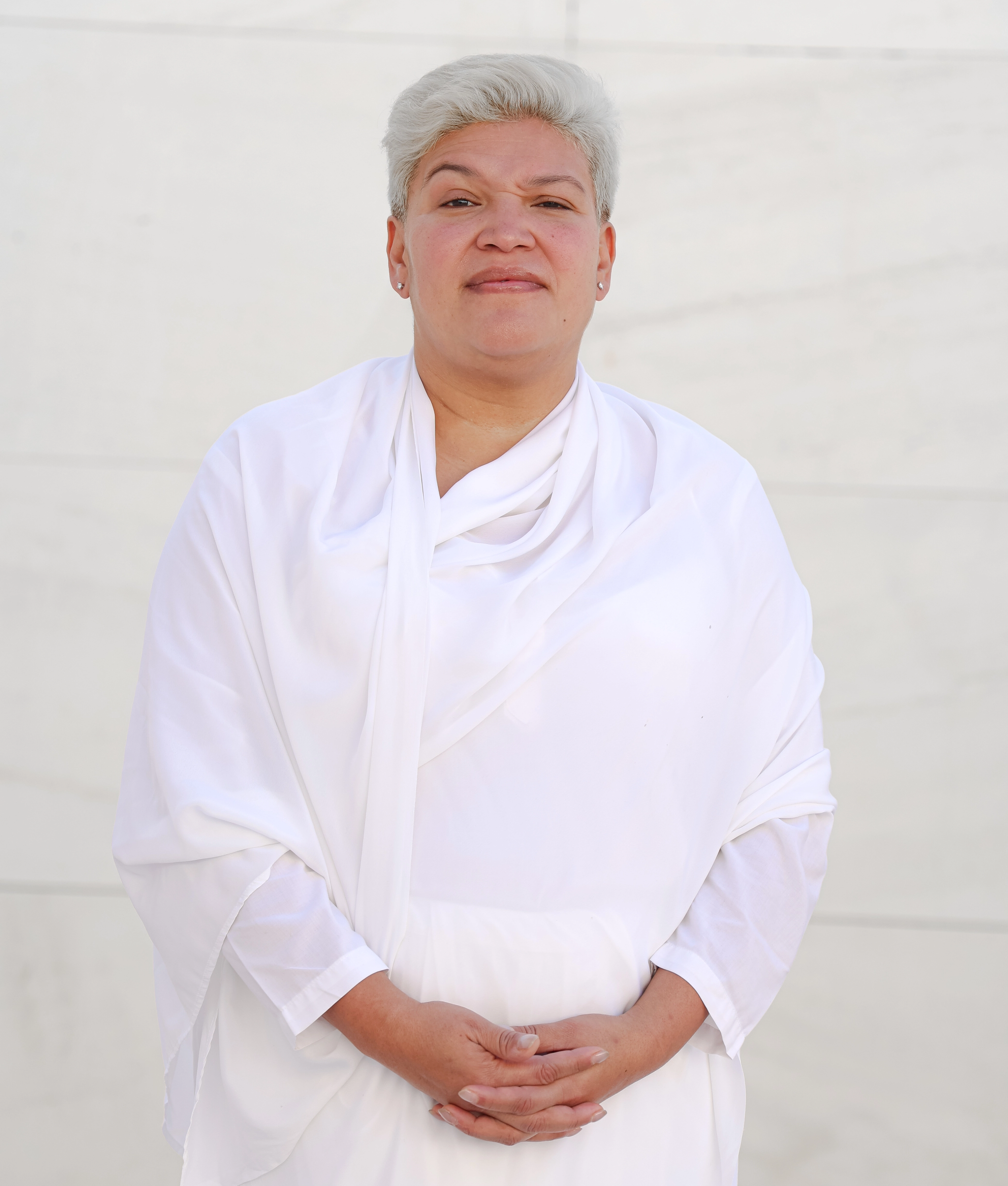 Persona de pie con cabello corto y plateado, vestida con una túnica blanca, sonriendo ligeramente frente a un fondo claro.