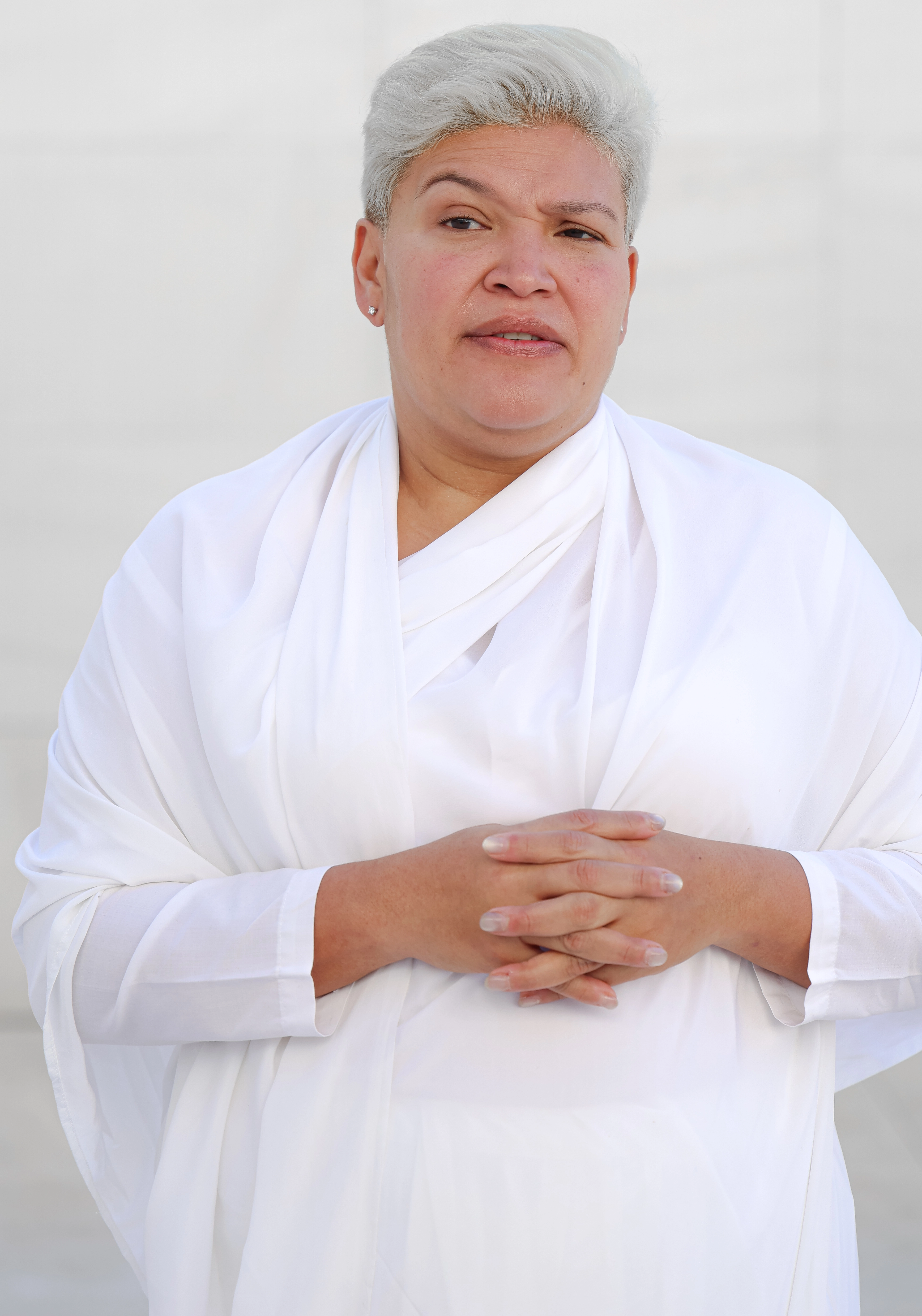 Una mujer con cabello corto y plateado, vestida con una túnica blanca, de pie con las manos entrelazadas frente a un fondo claro.