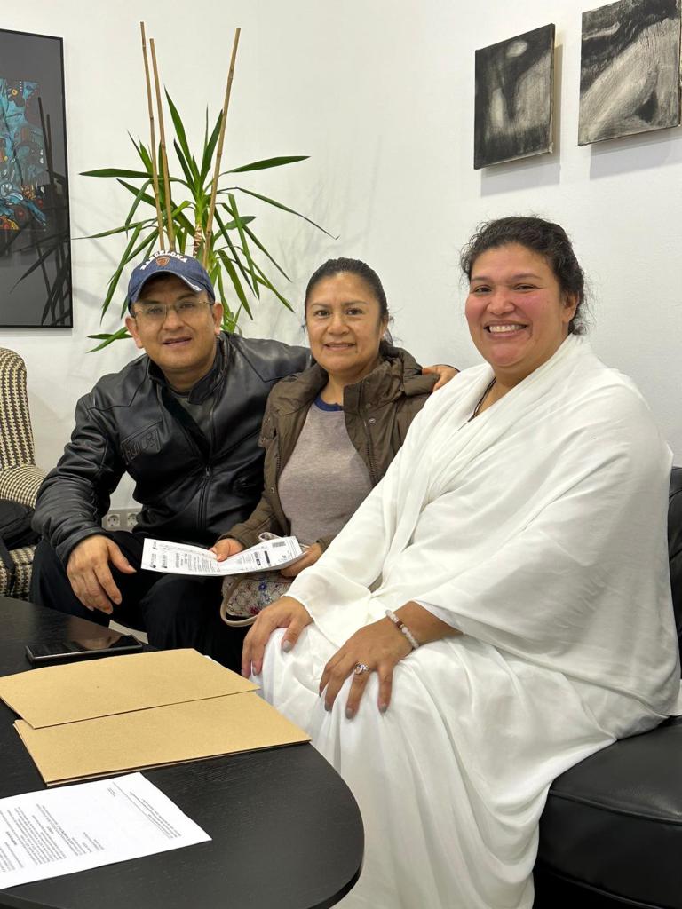 Tres personas sentadas en un sofá, sonriendo. Hay un hombre a la izquierda, una mujer en el centro y otra mujer a la derecha. En el fondo, plantas y cuadros decorativos.