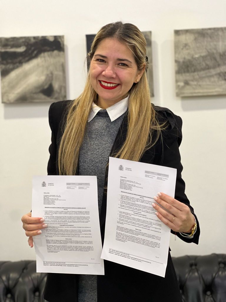 Mujer sonriente sosteniendo dos documentos en un entorno interior, con una pared blanca y una silla de cuero negro al fondo.