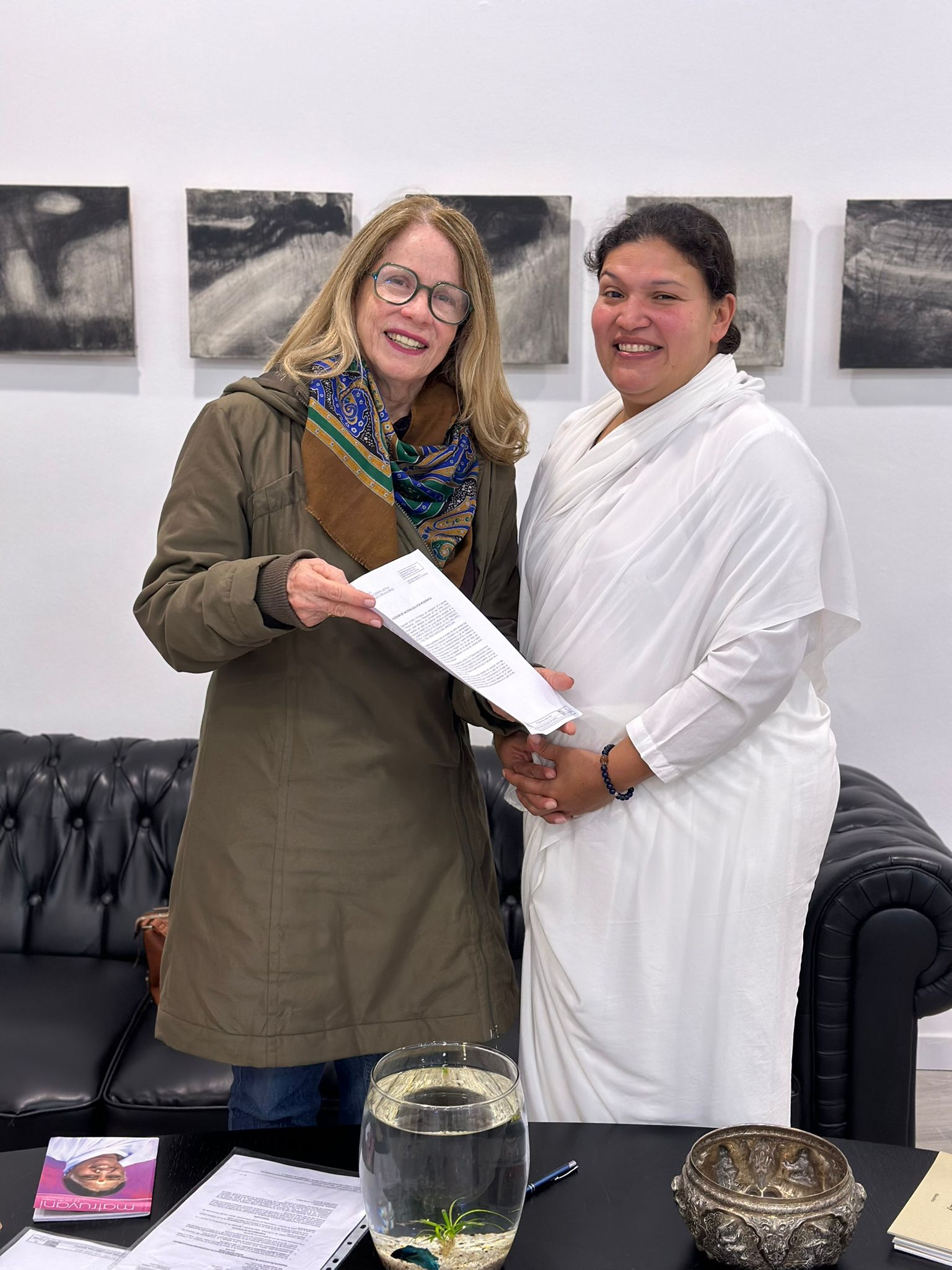 Dos mujeres sonrientes sosteniendo un documento en un entorno moderno, con pinturas abstractas en la pared y una mesa con un pez en un acuario.